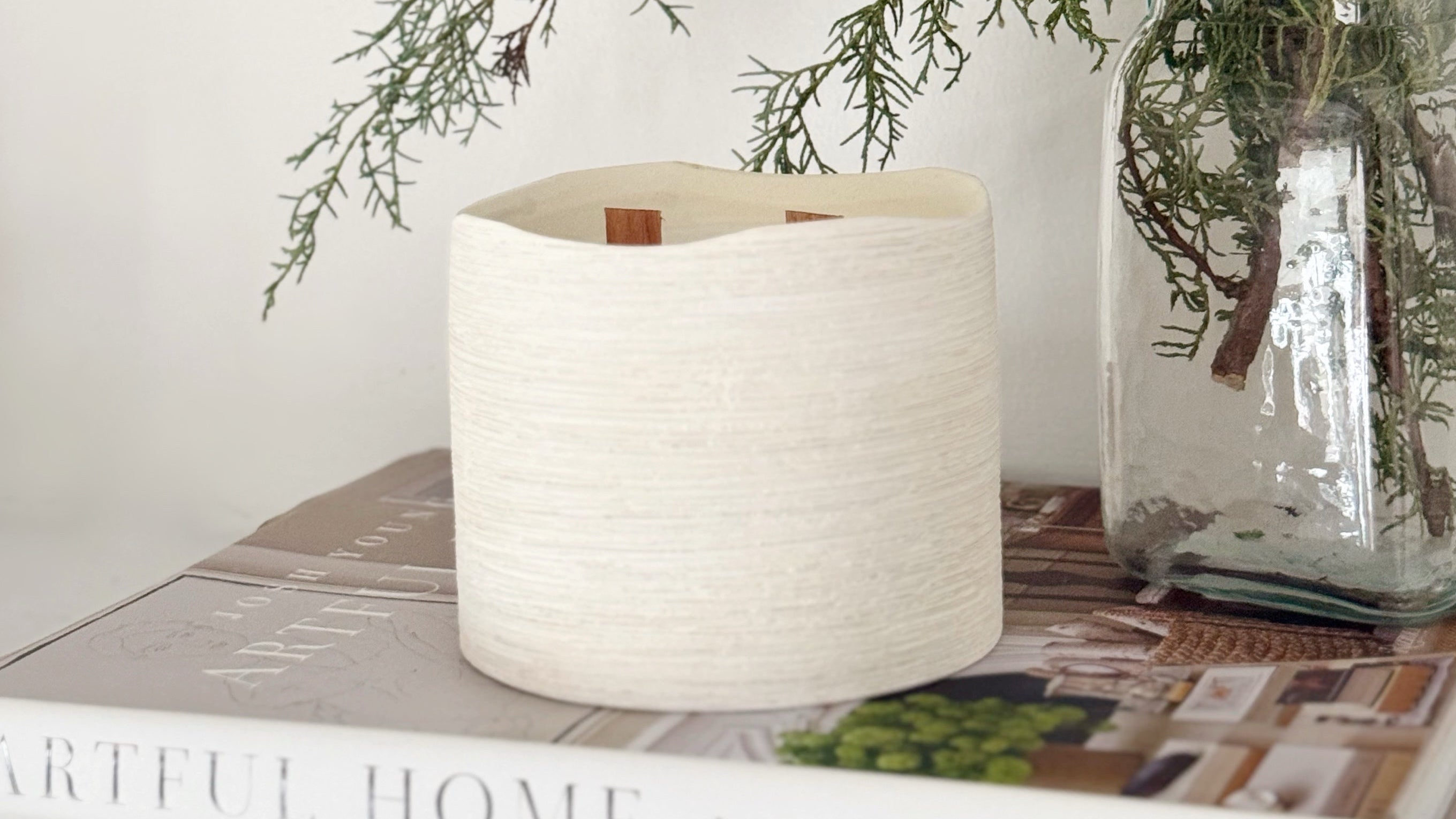 White textured candle and glass vase with greenery on a surface