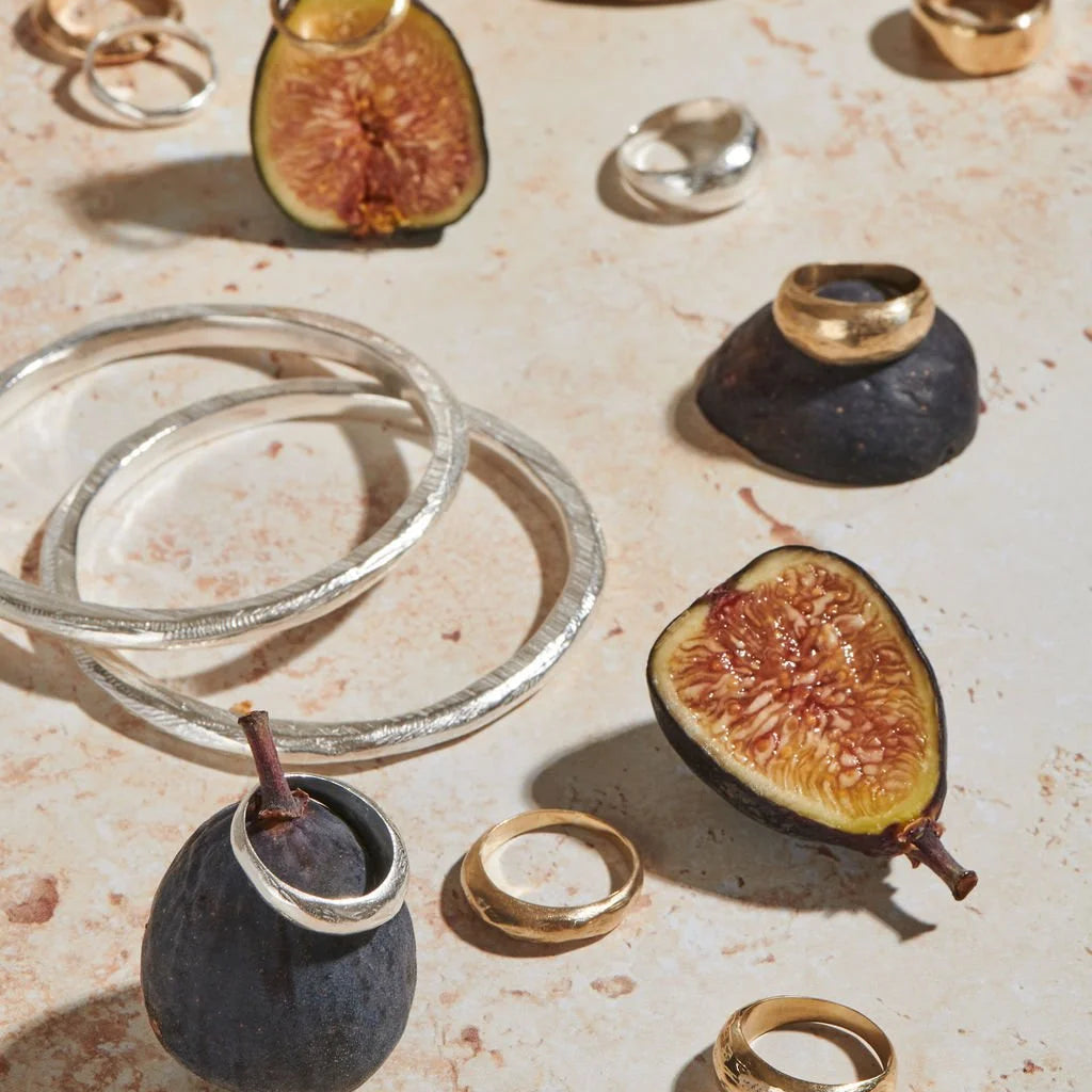 A styled flat-lay of mixed metal rings and bangles displayed on a textured beige stone surface, interspersed with fig halves and dark stone props.